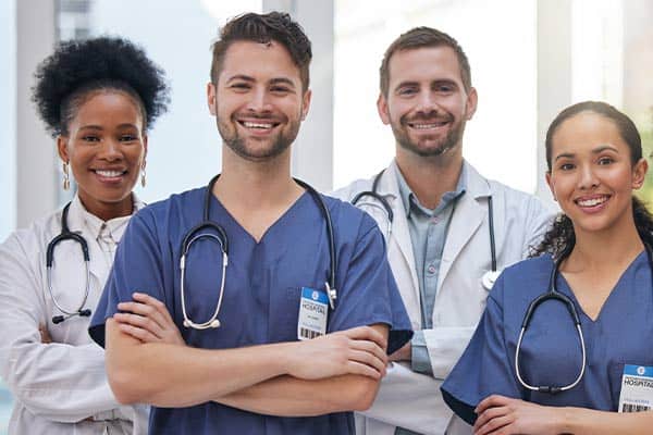 Four smiling primary care doctors wearing stethoscopes, with two in blue scrubs and two in white coats